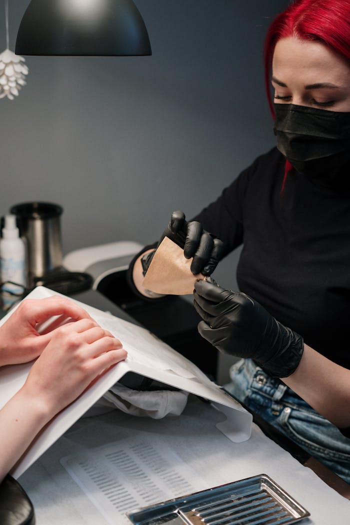 Nail technician with red hair applying nail art in a salon, wearing black gloves and mask.